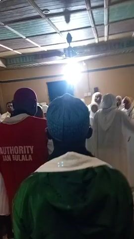 The video shows a gathering of people in what appears to be an indoor space. Several individuals are wearing white clothing, some with head coverings. There are decorative elements on the walls, including framed pictures and possibly flags or symbols. The lighting suggests an interior setting with overhead lights and possibly natural light coming from windows. The camera movement is shaky and sometimes blurry, indicating it was likely filmed by someone present in the crowd. The audio contains what sounds like chanting or speaking in a language that is not English. Some individuals in the foreground are looking away from the camera, while others are facing it. The atmosphere appears to be one of a religious or ceremonial gathering.
