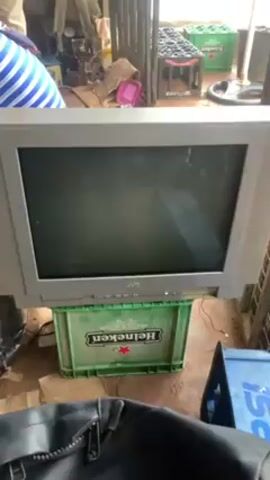 A JVC CRT television sits on a green Heineken crate, with a blinking light indicating it's on. A black leather jacket is partially visible in the foreground. To the right, there's a blue Pepsi crate. The background shows a cluttered indoor space with more crates, miscellaneous items, and sunlight streaming in. The TV screen is dark and inactive.