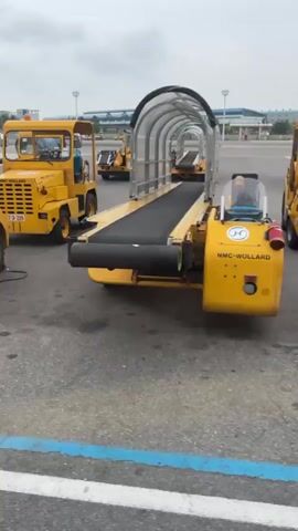 The video shows a series of yellow airport tugs parked on an asphalt surface, likely an airport tarmac. The tugs are small, utilitarian vehicles with open-air cabs. The foreground tug has the number 2-3-287 visible on its front. In the middle ground, another tug is visible, and further in the distance, a third tug is also present. The tugs appear to be lined up, suggesting they are parked or waiting for use. The environment appears to be overcast, with a building visible in the background and a line of blue and white paint on the asphalt in the foreground. The overall impression is one of an operational area at an airport, with service vehicles positioned and ready.