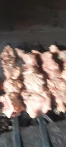 The video shows pieces of meat on skewers being grilled over coals. The meat appears to be chicken or lamb, and it is being cooked on a metal grill. Smoke is rising from the coals and the meat. A green object, possibly a brush or spatula, is briefly seen in the lower right corner of the frame, seemingly being used to turn or baste the meat. The lighting is dim, suggesting it might be nighttime or a poorly lit area. The overall scene depicts outdoor cooking.