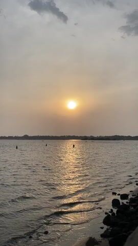 The video shows a sunset over a body of water with people wading in it. The sun is low in the sky, creating a golden reflection on the water's surface. There are a few individuals standing in the water at varying distances from the shore. The shoreline is visible in the foreground, consisting of sand and dark rocks. In the background, across the body of water, a line of trees can be seen, indicating a distant shore. The sky is overcast with some patches of lighter areas around the sun.