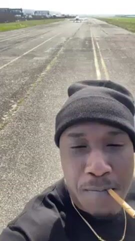 The video shows a person taking a selfie on an airport runway. The person is wearing a black beanie, a black shirt, and a gold chain. They are holding a cigar in their mouth. Behind them, a small white plane is on the runway. The runway is cracked and worn, with two yellow lines running down the middle. There are some buildings and greenery visible in the background under a sunny sky.