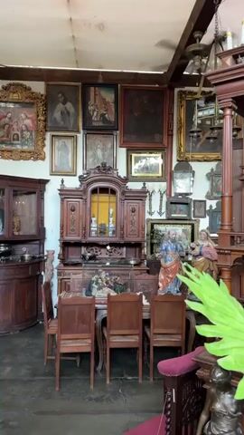 The video shows the interior of a shop or warehouse filled with antique furniture and artwork. The majority of the furniture is made of dark wood and is intricately carved. Pieces such as buffets, cabinets, chairs, and tables are visible. Numerous paintings, portraits, and religious iconography are hanging on the walls. There are also sculptures and decorative items scattered throughout the space. The ceiling is white and features chandeliers and ceiling fans. The overall impression is of a cluttered but well-maintained collection of antiques.