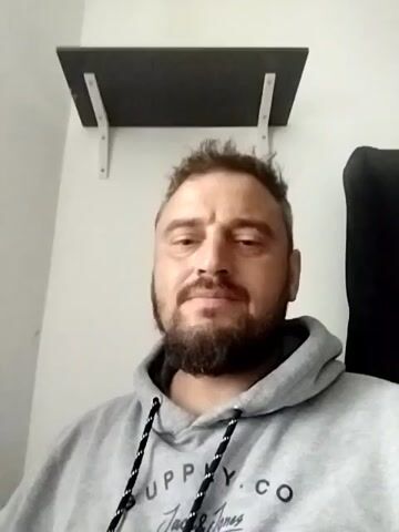 A man with a beard and a grey hoodie is in front of a white wall with a shelf above him. He smiles and waves at the camera.