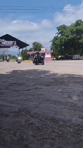The video shows a group of three motorcycles driving past. The motorcycles are driving on a slightly uneven dirt road. The sky is blue with some clouds. There is a large tree to the right. The motorcycles are moving from right to left in the frame. There are some buildings in the background. There is a stop sign in the middle of the road.