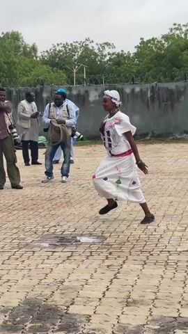 A woman in traditional African attire, including a white dress with colorful embroidery and a headscarf, dances in an outdoor courtyard paved with interlocking bricks. She is surrounded by several men, some of whom are holding cameras, suggesting they are photographers or journalists. The background features a grey concrete wall, a chain-link fence, and greenery. Several vehicles, including a white SUV, are parked nearby. The woman's movements are lively and expressive, with her arms extended and her body in motion. The overall atmosphere appears to be public and possibly ceremonial, given the presence of media and the traditional clothing.