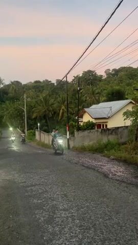 Two people ride a motorcycle down a wet, unpaved road lined with a concrete wall and lush green vegetation. A yellow house with a white roof sits behind the wall. Overhead, a tangle of power lines crosses the frame. The sky is a soft pink and orange, indicating sunset or sunrise. The motorcycle has its headlights on.