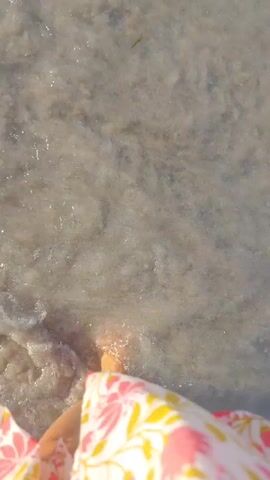A first-person perspective shot shows a person's legs and feet walking through shallow, clear water on a sandy bottom. The water is disturbed by their movement, creating ripples and splashes. The person is wearing a floral-patterned garment, which is visible at the bottom of the frame. The lighting suggests a bright, sunny day.