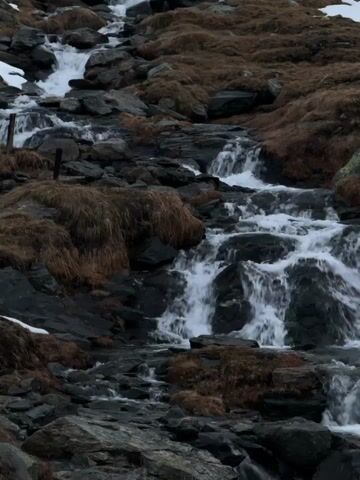 A stream cascades down a rocky, moss-covered slope in Poschiavo, Switzerland, creating multiple small waterfalls and rapids. The water is white and foamy as it rushes over dark grey and black rocks. Patches of dried, light brown grass cover parts of the slope, suggesting an alpine or high-altitude environment. Small remnants of snow are visible on the upper left side and scattered in other areas, indicating cool or cold weather conditions. The lighting is diffused, possibly overcast or during early morning/late afternoon. No people or visible text are present in the video.