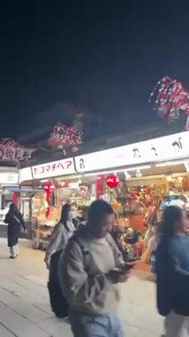 At night, a crowded pedestrian street is lined with shops lit with white lights that appear to sell gifts and souvenirs. Overhead, the street is decorated with red and white paper decorations.  People stroll along the street, some wearing traditional Japanese clothing. In the distance, a large temple structure with a red and dark-colored roof is visible. The sky above is a dark, clear blue.