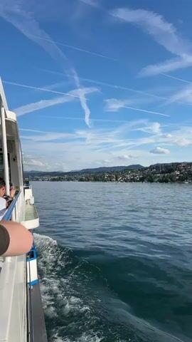 A scenic daytime video captures a boat cruising on a large body of water, likely a lake, under a bright blue sky crisscrossed with numerous airplane contrails. The boat, white with blue accents, is visible on the left side of the frame, creating a gentle wake behind it. A person's arm is visible in the immediate foreground, suggesting a passenger on the boat.

In the distance, a charming town with many houses and lush trees sprawls across a verdant hillside, stretching along the shoreline. The water surface is calm, reflecting the clear sky. The camera pans slightly from showing the side of the boat and people on board to revealing more of the expansive lake and the distant town. The overall setting is serene and picturesque, suggesting a leisurely boat trip on a sunny day in Kilchberg (ZH), Switzerland.