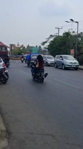 The video shows a street scene with vehicles and people. In the foreground, a white motorcycle is parked on the left side of the road. In the middle ground, a black pickup truck with a blue tarp is driving on the road, followed by a silver minivan and other vehicles. Several motorcycles are also seen on the road, with riders. The environment appears to be a street in a town or city, with buildings and trees visible in the background. The sky is overcast. The camera is moving, suggesting it was filmed from a moving vehicle.