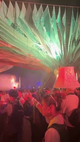 The video shows a crowded indoor event with people dancing and moving to music. A woman in the foreground is wearing a lamp shade on her head that reads "I LOVE LAMP". The lamp shade changes color with the lighting. The ceiling is decorated with strips of fabric in shades of red, white, and green. The environment is dimly lit, with smoke or fog in the air and stage lights visible in the background. People are wearing casual clothing and holding drinks.