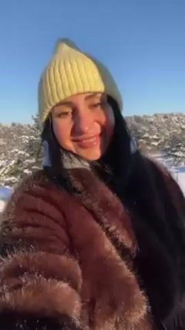 The video shows a woman, probably in her 40s, taking a selfie in a snowy environment. She is wearing a fur coat and a light green beanie. Behind her, there is a snowy landscape with trees and tracks in the snow. The sky is clear and blue. The woman smiles at the camera at the beginning. She briefly moves the camera angle. The lighting is bright, indicating a sunny day.