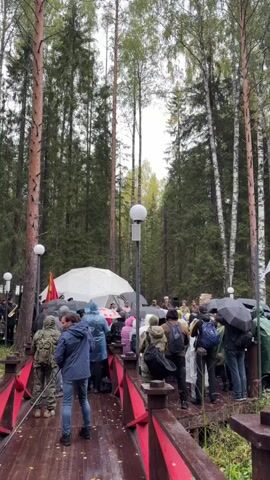 A group of people are gathered on a wooden bridge in a forest. Many are holding umbrellas, suggesting it is raining or has recently rained. The bridge has red railings with a decorative pattern. The people appear to be observing an event taking place under a large white tent in the background. Some individuals are wearing military-style camouflage clothing. There are trees with tall trunks and green foliage surrounding the scene. Flags are visible in the background. The atmosphere appears somber or formal, as indicated by the quietness and the focused attention of the onlookers.