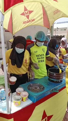 This media file captures an outdoor event, likely a food demonstration or promotional stand, in Tarauni, Nigeria. The scene is brightly lit, suggesting daytime, and the weather appears warm and sunny.

The central focus is a food preparation station under a large, yellow and red branded umbrella advertising "MAGGI". A man wearing a light blue hairnet, face mask, and gloves is actively cooking in a pot on a portable gas stove. He's stirring the contents with a yellow utensil. He wears a neon yellow t-shirt with black sleeves and black text on the front.

Several other individuals are present. To the left, a woman in a black hijab and yellow t-shirt, also wearing gloves and a face mask, looks down at the counter. In the background, a person in a red shirt with "BRISCO FOODS" printed on it is visible. To the right of the cook, a woman in a white hijab and yellow apron is holding something in her hand, possibly giving instructions or a product. Another woman in a yellow t-shirt and a black visor, with a long braided ponytail, is facing the cook and holding up a phone, appearing to be recording or taking pictures.

The counter is stocked with several individual serving cups of what appears to be instant noodles or similar food items, along with a bottle of water. The branding on the umbrella and the counter, featuring the "MAGGI" logo and stars, clearly indicates a promotional activity for the food brand. The overall atmosphere seems busy and engaging, with people actively participating and observing the food preparation. The presence of food branding, cooking demonstrations, and promotional materials suggests a marketing event designed to attract and engage consumers.