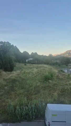 The video shows an outdoor scene with a clear blue sky. In the foreground, a white van is parked. Beyond the van, a grassy area slopes upwards towards a wooded hill. A few scattered trees are visible on the hillside, and in the distance, rolling hills under a bright sky. The video pans slightly to the right, revealing more of the landscape, including some buildings and structures among the trees.