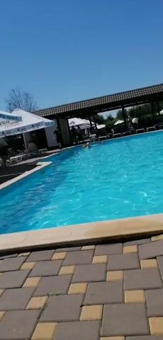 A clear, sunny day at a poolside resort. The camera pans across a bright blue swimming pool, revealing several people lounging on chairs under large white umbrellas. Some individuals are in the pool, splashing and playing, while others are relaxing by the water's edge. The ground is paved with a mosaic of brown and beige tiles. In the background, a building with a dark roof and a covered patio area can be seen, along with lush green trees. The umbrellas are branded with a logo and text that appears to be in Cyrillic. The overall atmosphere is one of leisure and enjoyment on a warm summer day.