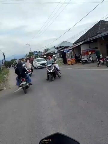 A street scene with moderate traffic. A white van is visible in the distance, along with several motorcycles. The motorcycles are carrying one or two people each. The environment appears to be a commercial area with buildings lining the side of the road. The sky is overcast.