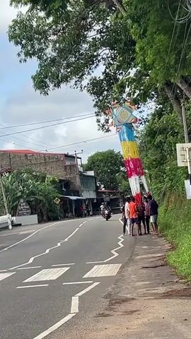 The video shows a street scene in Sri Lanka.  A large, colorful star-shaped decoration hangs over the road.  A group of five people stand on the shoulder of the road, near the decoration.  Two people are riding a motorcycle down the road toward the camera.  Buildings are visible on the left side of the road, and trees and a hillside are visible on the right. The sky is overcast.