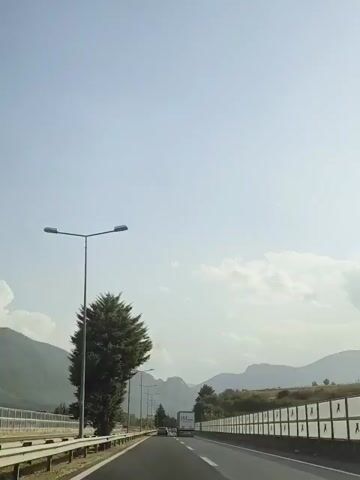 A daytime, first-person perspective video captures a drive along a highway. The camera is mounted to a vehicle, facing forward. A large, evergreen tree stands prominently to the left, with its branches extending over the highway. Several streetlights line the left side of the road. In the distance, mountains form a backdrop, with a hazy sky above. A white truck is visible in the right lane of the highway. The road is paved, with white lines marking the lanes. The right side of the highway features a sound barrier with a repeating triangular pattern. The overall environment appears to be a rural or semi-rural highway setting.