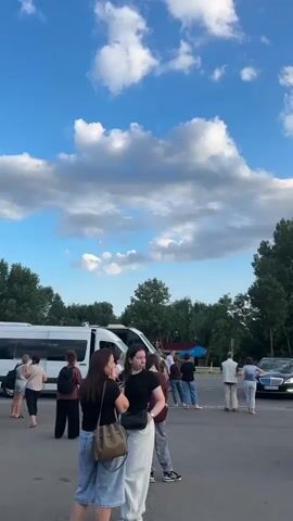 The video opens with a wide shot of a parking lot with several vehicles, including white vans, a black Mercedes sedan, and a teal van with its doors open. A group of people are standing and talking near the white vans. The sky is blue with scattered clouds. The camera then pans to the right, showing more of the parking lot and a border crossing checkpoint with signs and barriers. The teal van is parked in the foreground, with its open doors revealing the interior. The black Mercedes sedan is driving through the checkpoint. The video ends with a shot of the border crossing checkpoint, with trees and the sky in the background.
