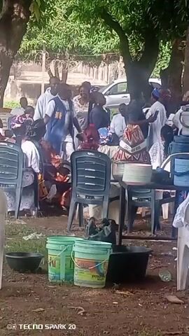 A group of people are gathered outdoors, possibly for an event. The scene is set under trees with some foliage visible. In the foreground, there are several plastic buckets and bowls on the ground. The people are dressed in various attire, some in traditional clothing. Some individuals are standing and interacting, while others are seated on plastic chairs around tables. There are stacks of bowls or containers on the tables. A vehicle is partially visible in the background. The ground appears to be dirt or gravel.