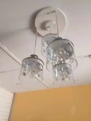 The video begins with a shot of a ceiling light fixture with four glass shades. The camera then pans down and to the left, showing a plant in a white pot next to a stack of white plates on the floor. There are also some blue pipes stacked near the plant. The background walls are white with a brick-like texture and yellow. There is a fan and a gold-colored frame in the background.