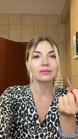 A woman with blonde-highlighted hair tied back is applying pink lipstick. She is wearing a black and white leopard print dress. The background appears to be a bathroom with beige tiled walls. She is holding a lipstick applicator to her lips and appears to be concentrating on her application. She is wearing bracelets on her left wrist.