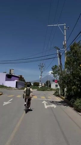 A person rides a bicycle down a street under a clear blue sky. The person is wearing a backpack and a hat. The street has yellow crosswalks and traffic arrows painted on the asphalt. There are utility poles with many wires running across the sky. In the distance, a mountain with a plume of smoke or clouds erupts from its peak. On the right side of the street, there is a red and white stop sign with "1x1" written on it. Buildings and vegetation are visible on both sides of the street.