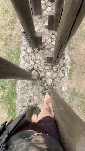 A first-person perspective video shows a person walking barefoot across an obstacle course made of wooden logs. The person is wearing dark shorts and a black bag is visible on their left hip. The ground of the obstacle course is made of cobblestones. The background shows grass and dirt. The person's feet are visible as they step from one log to another.