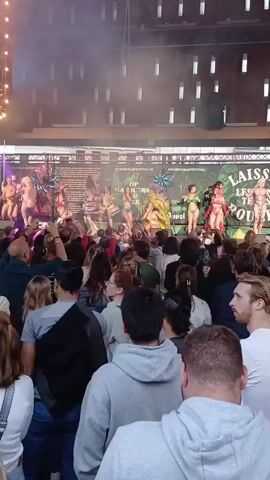 A crowd of people are watching a performance on a stage. The performers are dressed in revealing costumes, some with elaborate headpieces. There are lights and smoke effects on the stage. The background appears to be a building with windows. The audience is diverse in age and gender, and many are holding up phones to record the event.