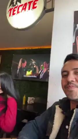 Two people are at a restaurant. A man in a denim jacket turns toward the camera and smiles. The woman next to him, wearing a red jacket and sunglasses, holds up a bottle of dark beer. There's a TV on the wall showing a program, plus decorative elements like a neon light, artificial greenery, and framed pictures. The restaurant has dark furniture.