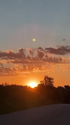 The video shows a sunset with the sun low on the horizon, casting a warm orange glow across the sky. There are scattered clouds, some illuminated by the sun, creating a picturesque scene. Silhouetted against the sunset are trees and a faint outline of power lines. The camera appears to be moving slightly, perhaps from a vehicle, as the perspective of the trees and horizon shifts. The overall environment is rural or natural, with no people or clear man-made structures visible other than the power lines. The lighting is characteristic of twilight.