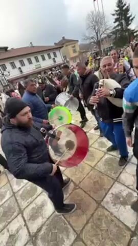 A wide-angle, low-angle shot captures a lively outdoor scene with several people playing music and celebrating. The primary focus is on three men in the foreground: one beating a large red drum, another with a neon green drum, and a third playing a bagpipe. The man with the red drum wears a black beanie and a dark jacket and is intently focused on his playing. Behind them, a crowd of people is gathered, with some wearing colorful traditional costumes. The architecture in the background suggests a town square or public space, with buildings featuring light-colored facades and traditional designs. The atmosphere appears festive and energetic, with the musical performance acting as the central point of attraction. The sky is overcast, indicating a cloudy day.