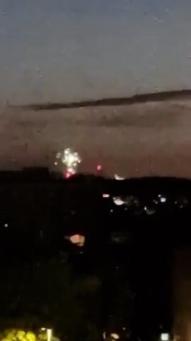 This is a video recording of fireworks being set off over a dark, silhouetted landscape at dusk. The sky is a dusky blue with some clouds. The fireworks explode in a variety of shapes and colors, including bright white and colorful bursts. There are also smaller red lights visible in the distance, which could be other fireworks or lights from buildings. The video is shaky and the image is somewhat grainy, suggesting it was filmed handheld and possibly from a distance.