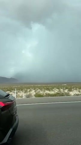 A green Lexus UX is shown from the side as it drives down a paved road in a desert landscape. The sky above is overcast with dark storm clouds. In the distance, mountains can be seen through a hazy sky. A small white building and two yellow construction vehicles are visible in the far distance.