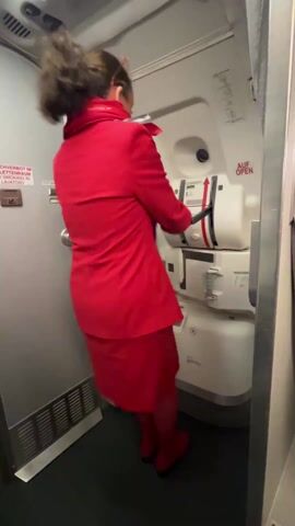 The video shows a woman wearing a red skirt and blazer with matching tights and shoes opening an airplane door.  The interior of the plane is visible, including a door labeled "smoking in lavatory" and overhead compartments. The door is opened to reveal an apron and a person in a yellow vest.