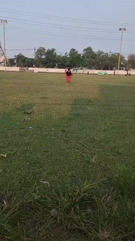A person wearing a red dress and a black hijab runs across a grassy field. There are stadium lights and trees in the background.