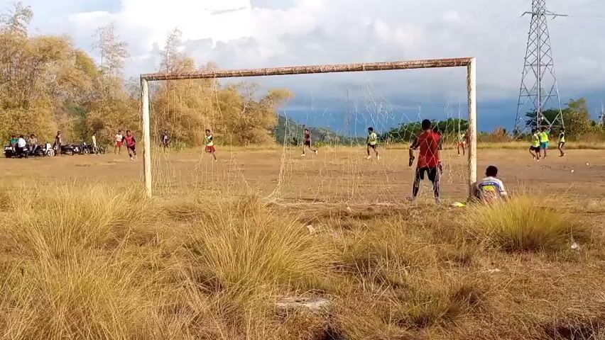 The video captures a group of young people playing soccer on a dry, grassy field. In the foreground, tall, golden grass partially obscures the view of the soccer game. A large, weathered soccer goal with a net dominates the center of the frame. Several players are visible on the field, wearing a mix of colorful shirts and shorts. Some are actively engaged in the game, chasing or kicking a ball, while others appear to be standing or observing. In the background, a line of motorcycles is parked, and a cluster of trees, some appearing dry and autumnal, forms the backdrop. A power line tower is also visible in the distance. The sky is partly cloudy. The overall impression is of an informal soccer match taking place in a rural or semi-rural outdoor setting.