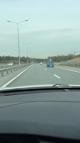 The video shows a car’s dashboard view while driving on a multi-lane highway under an overcast sky. Two trucks and several cars are visible on the road ahead, driving in the same direction. The highway has guardrails on both sides and is lined with tall streetlights. Trees can be seen in the background. The car’s windshield wipers are not in use.