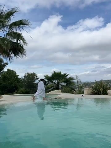 A person wearing a white robe stands with their back to the camera, at the edge of a light blue swimming pool. The person's legs are submerged in the water. Palm trees and lush green vegetation are visible in the background, along with a hilly landscape under a cloudy sky. The scene appears to be a resort or spa.
