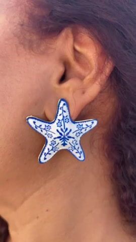 The video begins with a close-up of a woman's ear, adorned with a star-shaped earring featuring a blue and white floral pattern. The background is blurred and appears to be outdoors with bright sunlight. The video then transitions to a medium shot of the same woman, who has red curly hair and is wearing a blue dress with ruffled straps and a large bow in the back. She is standing on a balcony or rooftop overlooking a white village with buildings cascading down a hillside towards the sea. The woman is holding a white tissue to her nose and appears to be wiping it. She looks to her left and smiles. The sun is shining brightly, creating lens flare in the video. The final shot is a wide aerial view of the same white village and the blue sea with boats in the distance. The overall impression is a scenic, sunny location, likely in Greece.