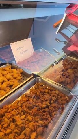 A food display case with various fried food items is shown. A handwritten sign in the upper left corner reads "W***** fish & chips menu - Kibbeling + soup & snacks". The food items appear to be fish and chips, calamari, and possibly shrimp. In the upper part of the frame, there are some decorative pink lobsters attached to the ceiling. The overall environment appears to be a food truck or a casual eatery.