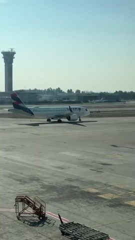 A LA TAM airplane is shown on a tarmac at an airport. The airplane has "LATAM" written on its side in blue letters and a logo with red, blue, and white colors on its tail. It is facing to the right and is on a paved surface with yellow markings. In the foreground, there are some industrial carts and a red staircase. In the background, there are buildings, trees, and a control tower. The sky is clear and blue, indicating a sunny day. The scene appears to be a typical airport operational area.