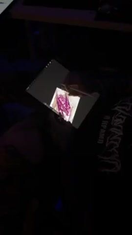 A person is sitting down in a dimly lit room, drawing on a tablet. The tablet is on a table, and the person is using a stylus to draw over an existing image on the screen, using pink lines. The person is wearing a black shirt.