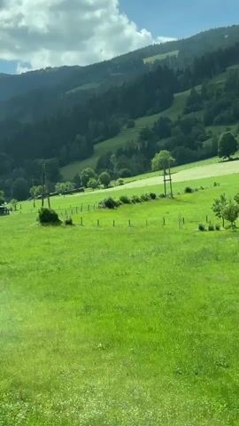 The video pans across a lush green meadow with rolling hills and forested mountains in the background. Several buildings, including a rustic wooden structure and a cluster of houses, are visible in the distance. A lamppost stands in the foreground, and power lines stretch across the landscape. The sky is partly cloudy.