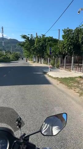 The video is a first-person perspective from a motorcycle or scooter on a sunny day. The road is paved, with yellow lines marking the lanes. On the right side of the road, there is a sidewalk and then vegetation and fences. On the left side, there are trees, utility poles, a cell tower, and distant mountains. The rider is approaching other vehicles on the road, including a motorcycle with a rider and a dark-colored car. The sky is clear and blue.