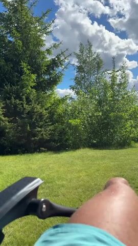 The video shows a first-person perspective of a person lying down on a lounge chair outdoors on a sunny day. The camera is pointed upwards, showing a bright blue sky with fluffy white clouds. In the foreground, the person's legs and feet are visible, along with a part of the lounge chair. Beyond the legs, a lush green lawn stretches out, with trees and bushes in the background. To the right, a low brick wall is visible. The overall scene depicts a relaxed outdoor setting.