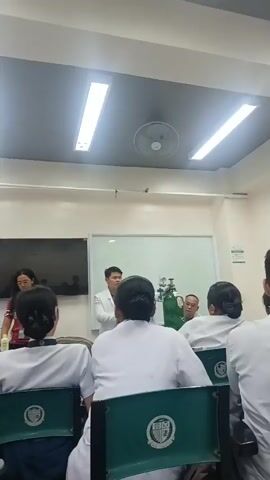 A group of students, some wearing white uniforms, are seated in a classroom facing a whiteboard and a large screen. Two people stand at the front of the room, with one person in a white lab coat addressing the students. Next to the person in the lab coat is a green oxygen tank with a regulator. The room is lit by overhead fluorescent lights, and a ceiling fan is visible. The students appear to be engaged in a lesson or presentation.
